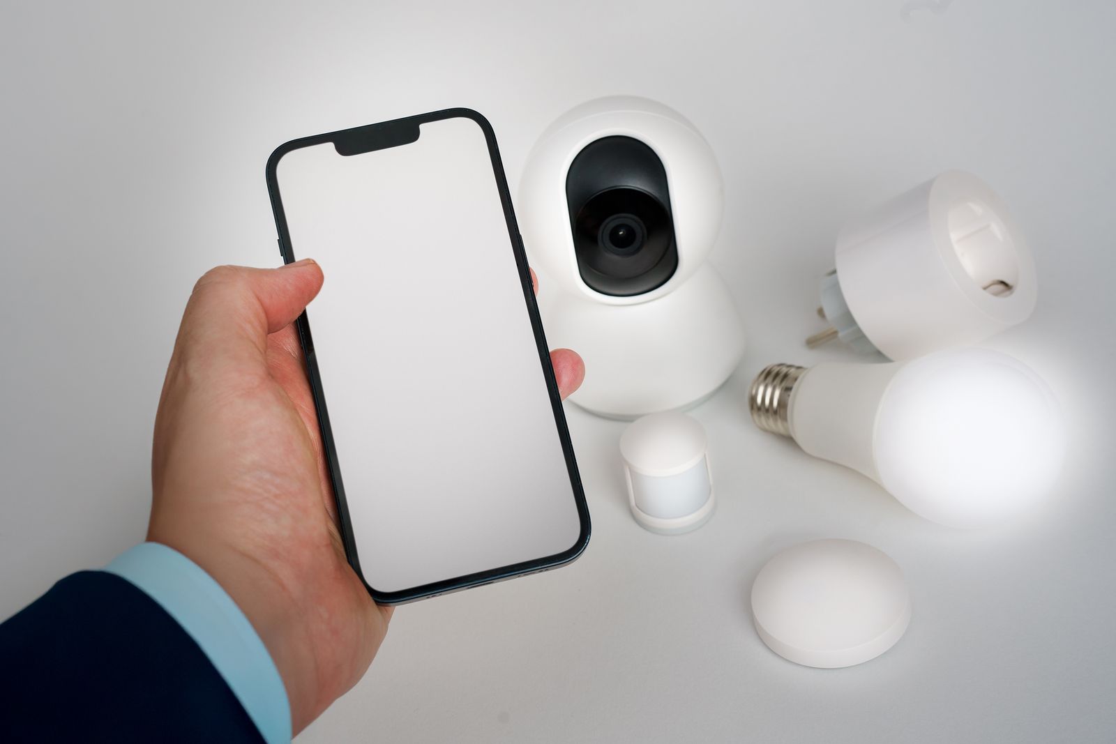 A hand holding a smartphone beside a smart camera, smart bulbs and sensors